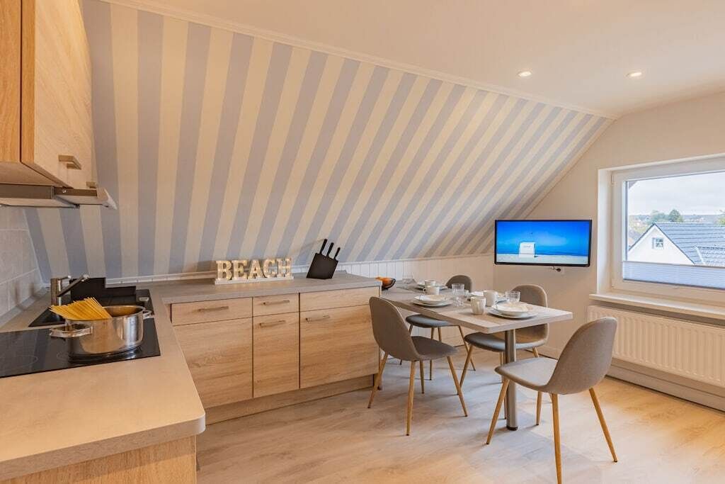 A modern kitchen with a dining area, a TV mounted on the wall, and a view of the outside through a large window.