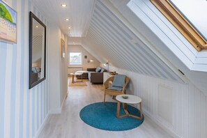 Una habitación con techo inclinado, un tapete azul redondo, una silla de mimbre, una pequeña mesa blanca y un cuadro en la pared.