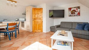Een compacte leefruimte met een kitchenette, een eethoek met blauwe stoelen, een gestreepte bank, een flatscreen-televisie en een schilderij aan de muur.