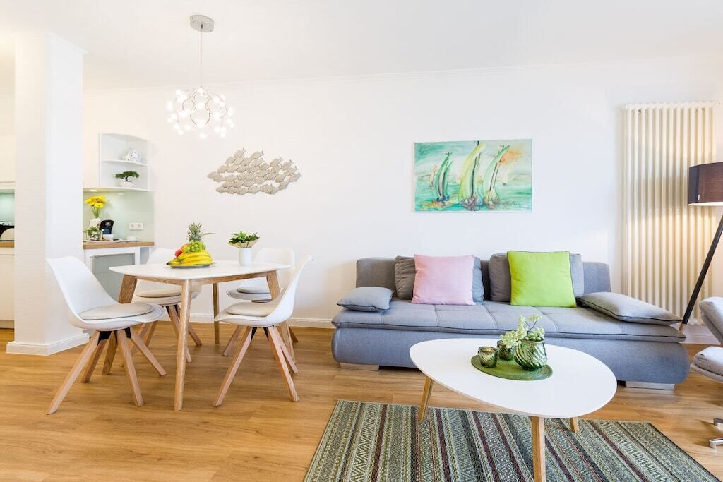 A modern living room with a grey sofa, white dining table, and chairs. There is a painting on the wall and a plant on the sofa.