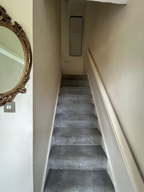 A carpeted staircase with a handrail, a wall-mounted light switch, and a mirror with an ornate frame. - Spacious 2BR Home | Attic Room, Parking & WiFi (Sunderland)