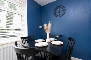 Un coin repas avec une table dressée pour quatre personnes, une horloge murale et une fenêtre avec des stores.