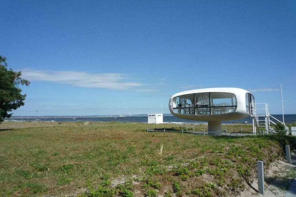 En modern, vit byggnad i kupolform med en glasfasad, belägen på en gräsyta med fri utsikt över havet i bakgrunden.