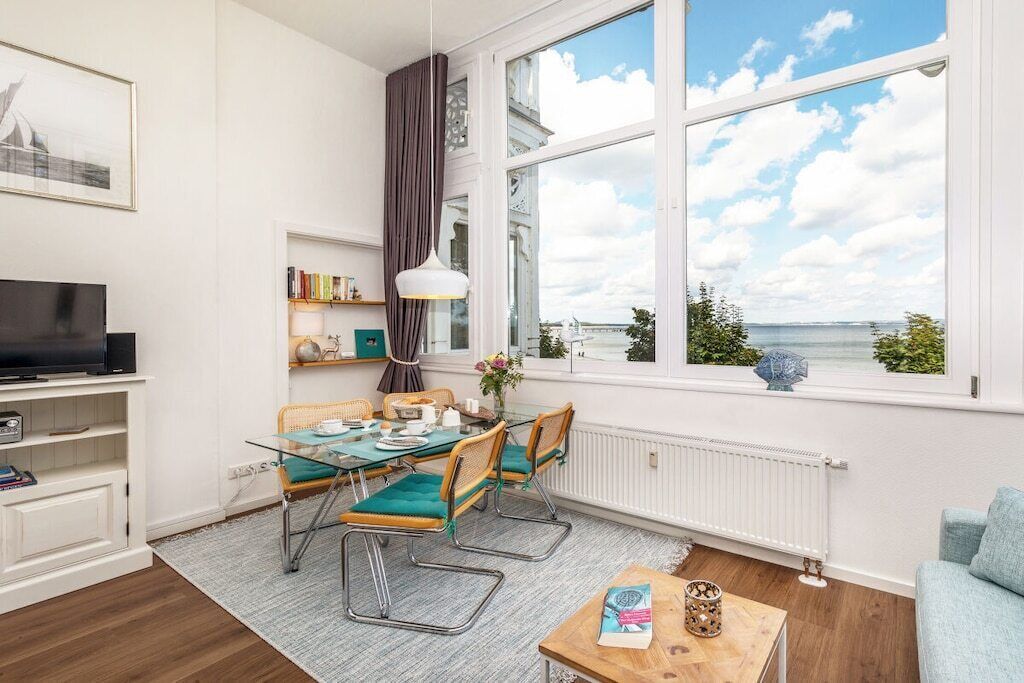 Una sala de estar moderna con televisión, mesa de comedor y sillas, un ventanal con vista al mar y un rincón de descanso acogedor.