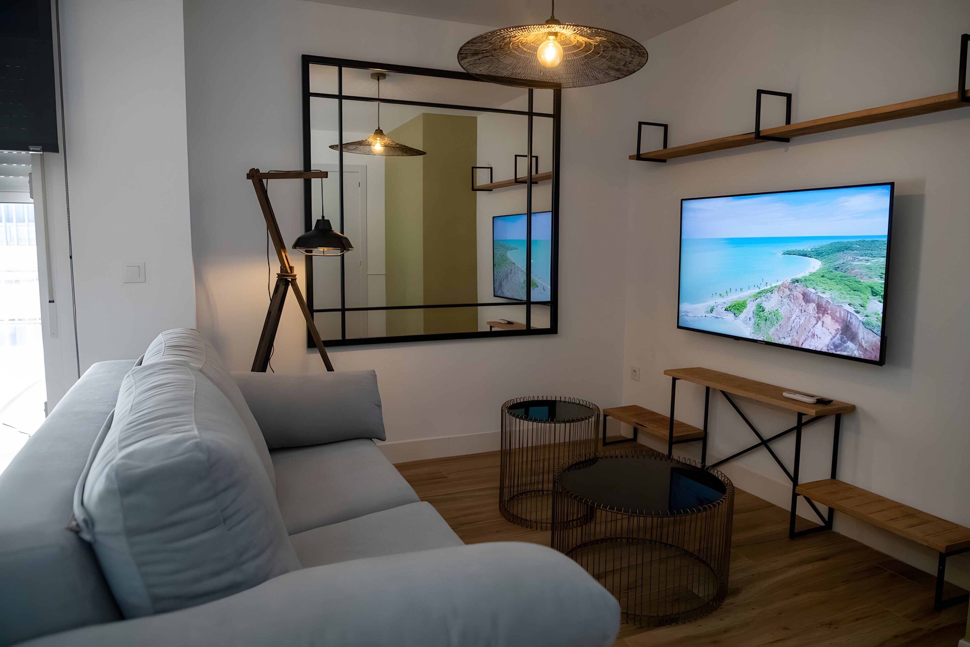 A modern living room with a white sofa, a large flat-screen TV, a floor lamp, a mirror, and a wooden coffee table.