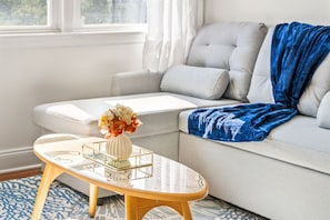 A modern living room with a sofa, a coffee table with a vase of flowers, and a blue blanket draped over the sofa.
