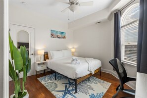 Une chambre d’hôtel avec un lit, des lampes de chevet, un ventilateur de plafond, une fenêtre avec des stores et une grande plante.
