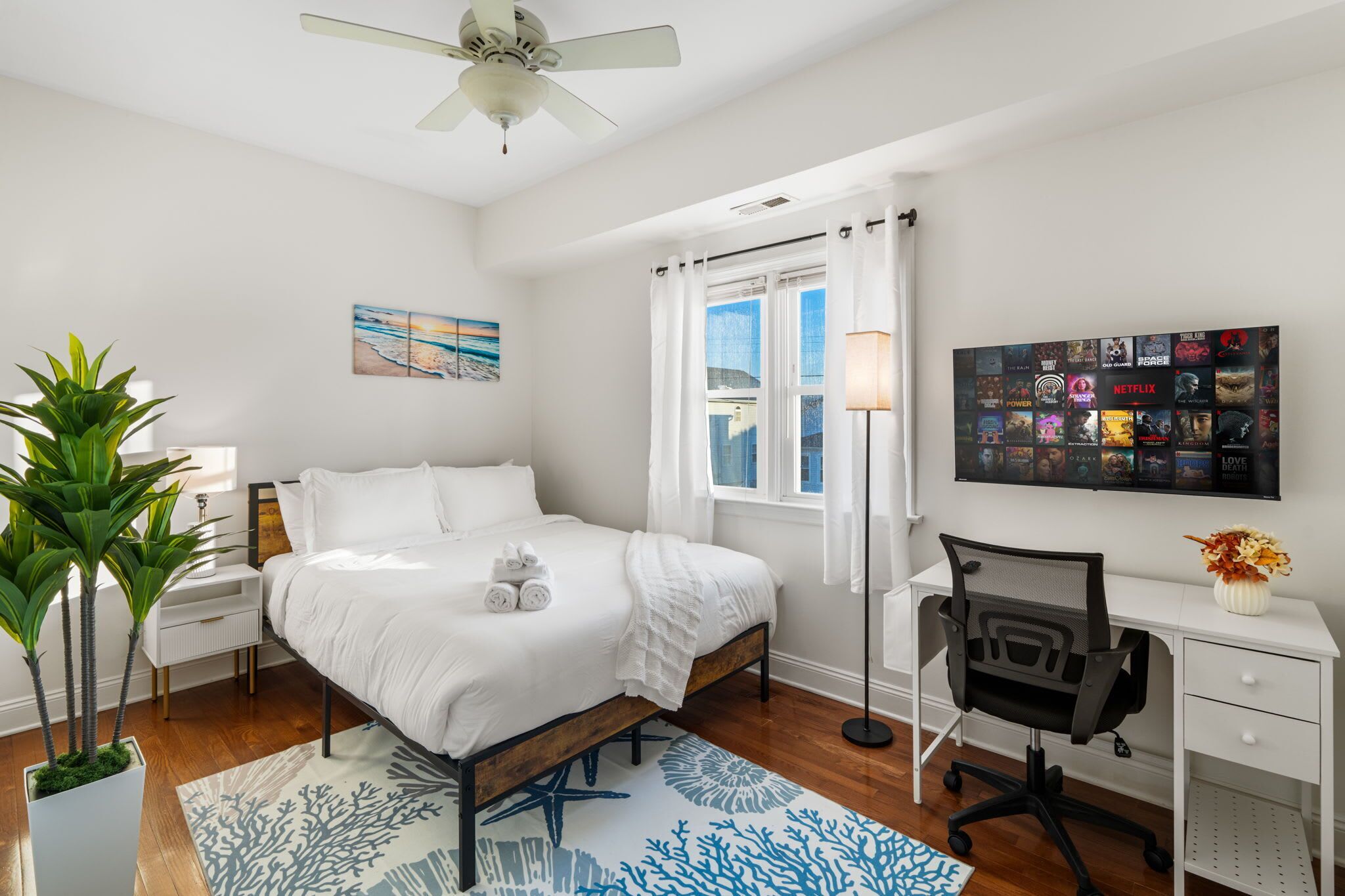 A modern bedroom with a bed, bedside tables, a desk, a chair, a potted plant, a painting, and a window with curtains.