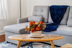Una sala de estar con un sofá gris, una manta azul y una mesita redonda con un centro decorativo.