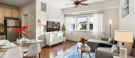 A modern living room with a dining area, a ceiling fan, a flat-screen TV, and a large window with curtains.