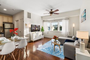 A modern living room with a dining area, a ceiling fan, a flat-screen TV, and a large window with curtains. - Breezy Beach Getaway, 3 Min Walk to Beach (Brigantine)