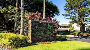 Ein Steinschild mit der Aufschrift 'Kapalua Golf Villas' und einem Schmetterlingslogo, umgeben von üppiger Vegetation und Bäumen.