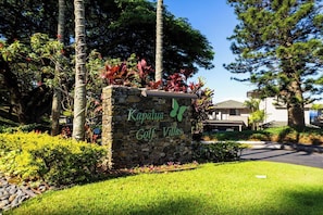 Un letrero de piedra con la inscripción 'Kapalua Golf Villas' y un logotipo de mariposa, rodeado de exuberante vegetación y árboles.