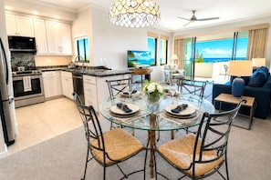 A modern kitchen with white cabinets, a dining area with a glass table and wicker chairs, and a living area with a view of the ocean. - Kapalua Golf Villas | 1-Bedroom Ocean View | KGV-23T1 by KBM (Lahaina)