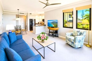 A modern living room with a blue sofa, a coffee table with flowers, a patterned armchair, a TV, and a kitchen in the background. - Kapalua Golf Villas | 1-Bedroom Ocean View | KGV-23T1 by KBM (Lahaina)