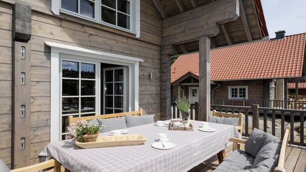 Una terraza de madera con una mesa lista para comer, rodeada de sillas de madera y un banco acolchado.