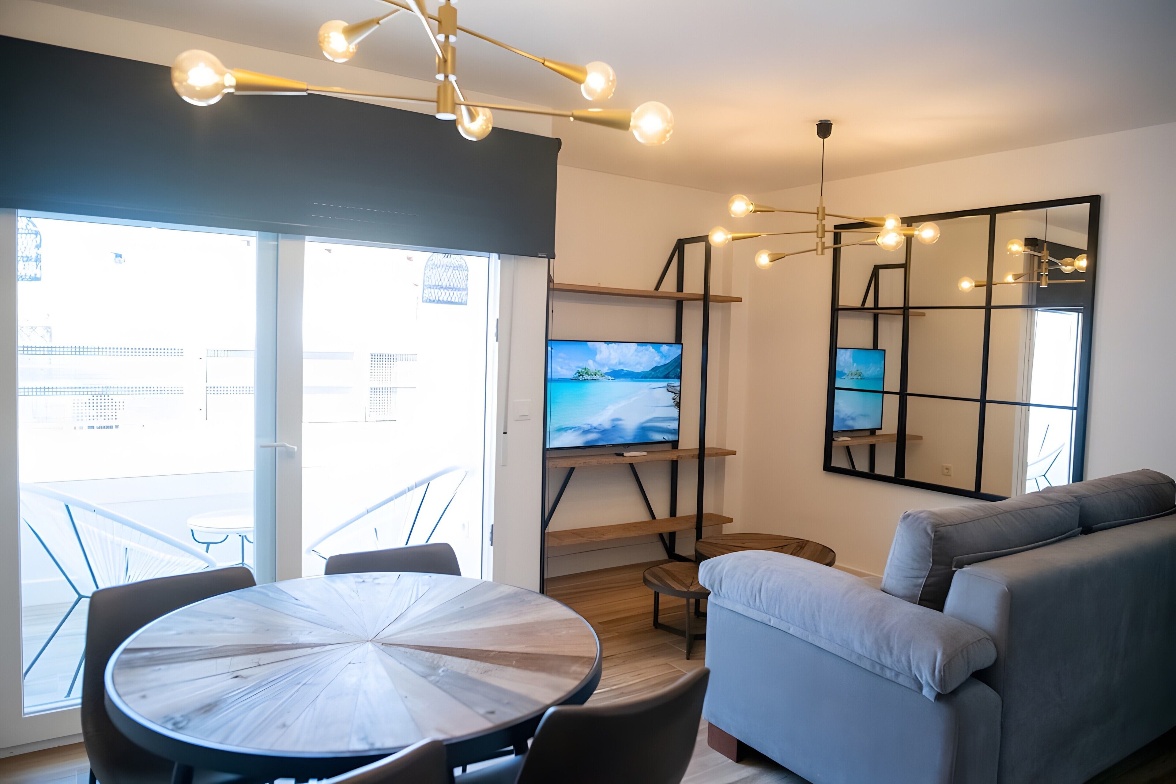 A modern living room with a round table, a grey sofa, a TV on a wooden shelf, and a large window with a view of buildings.