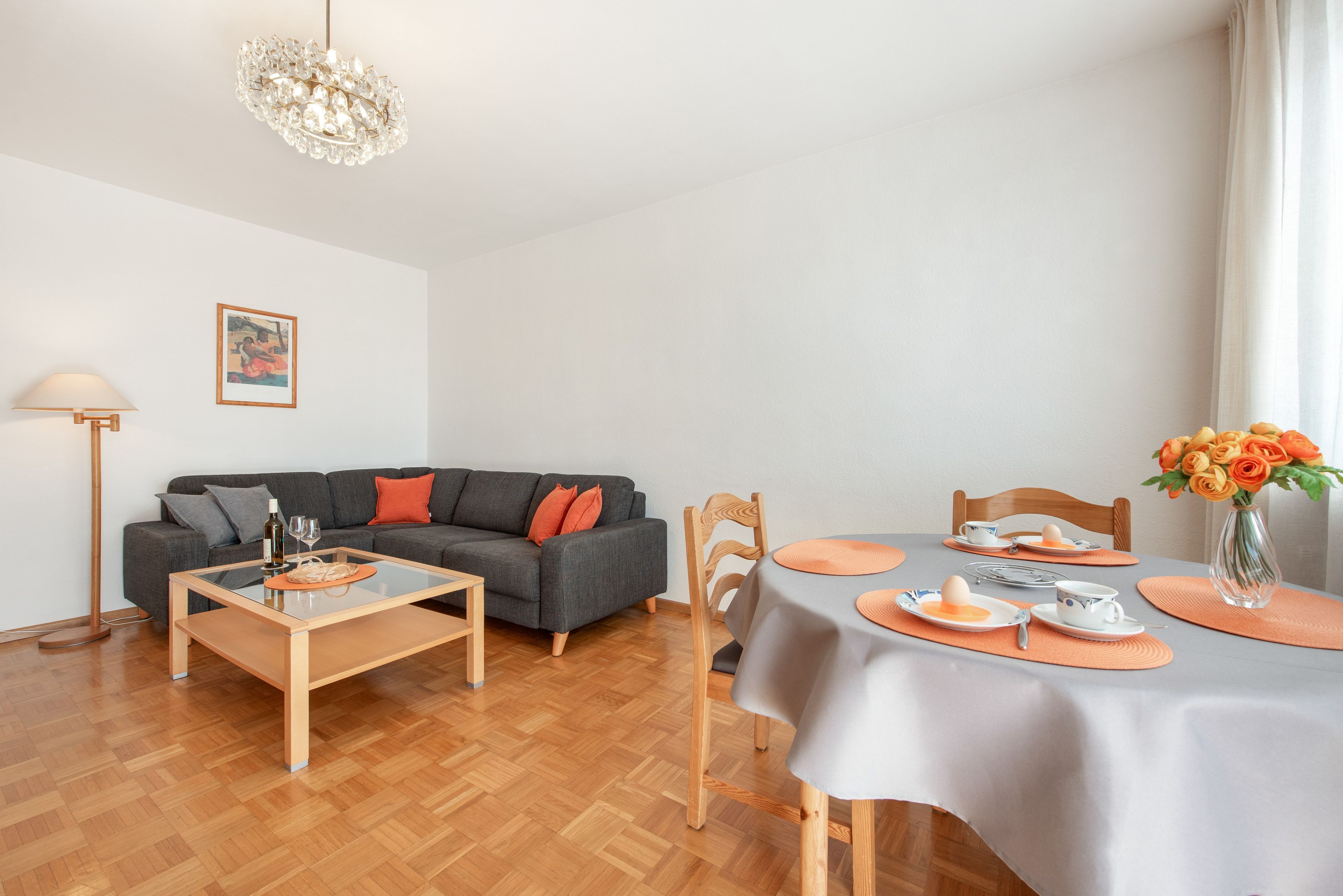 Ein modernes Wohnzimmer mit einem grauen Sofa, einem Esstisch für zwei Personen und einer Chandelüster.