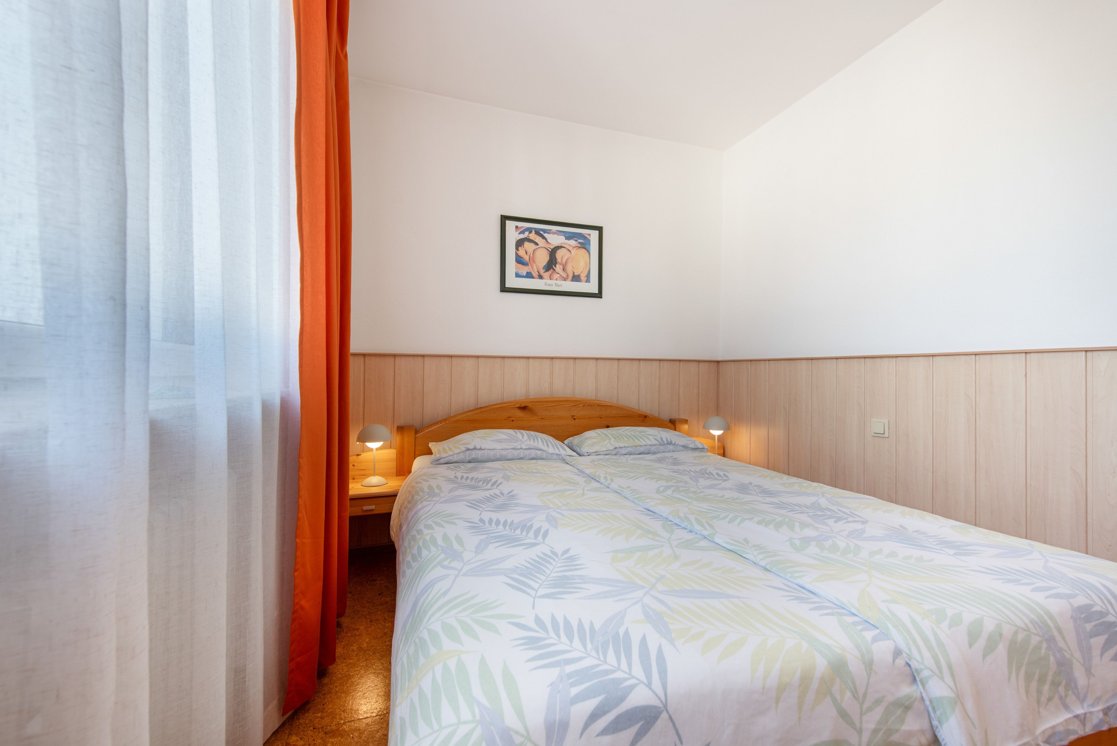Ein Schlafzimmer mit einem Bett, einem Nachttisch, einer Lampe, einem gerahmten Bild an der Wand und einem Fenster mit Vorhängen.
