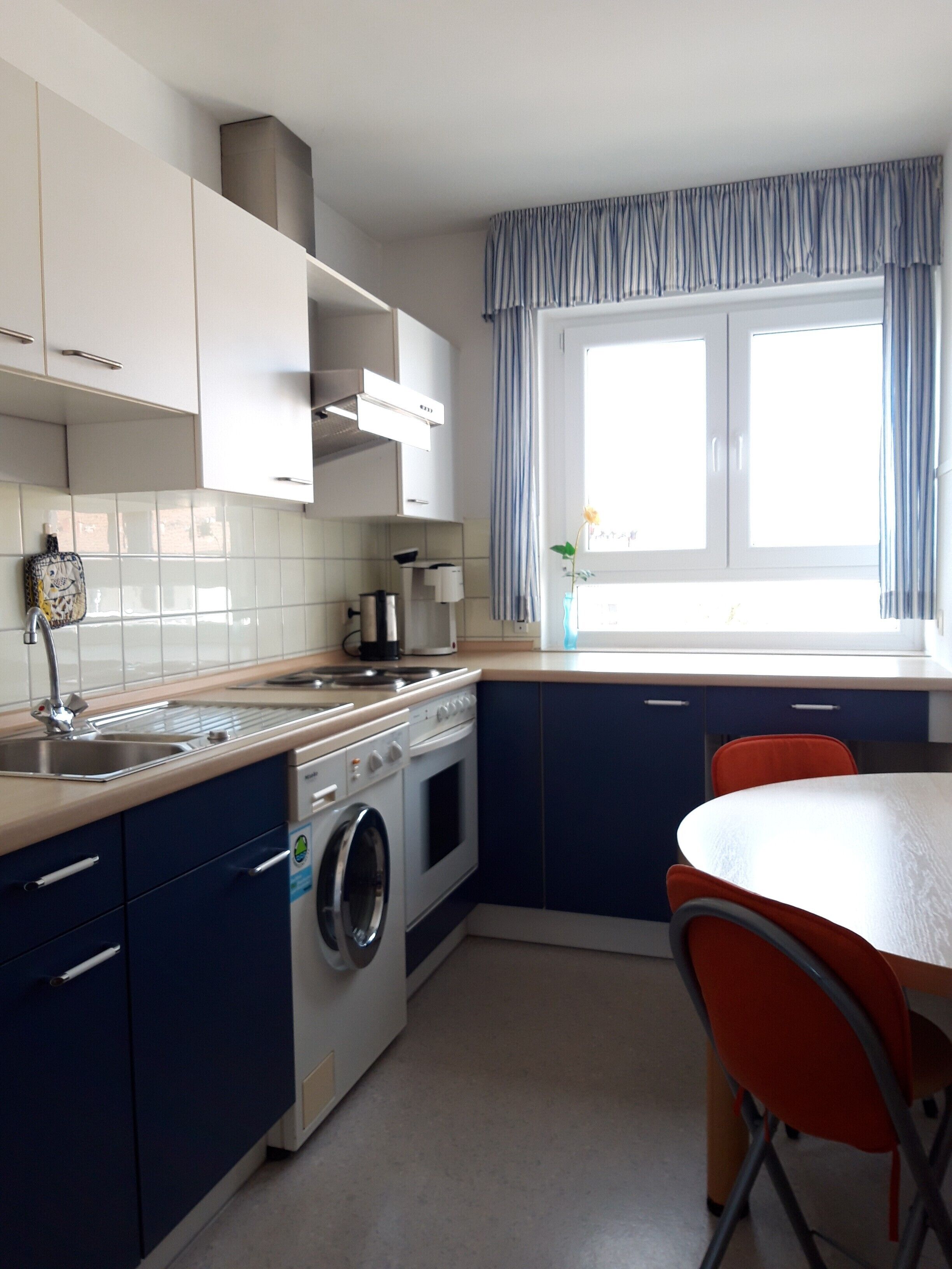 Une cuisine compacte équipée d’un lave-linge, d’une cuisinière et d’un évier. On y trouve une fenêtre avec des rideaux, ainsi qu’une table et des chaises.