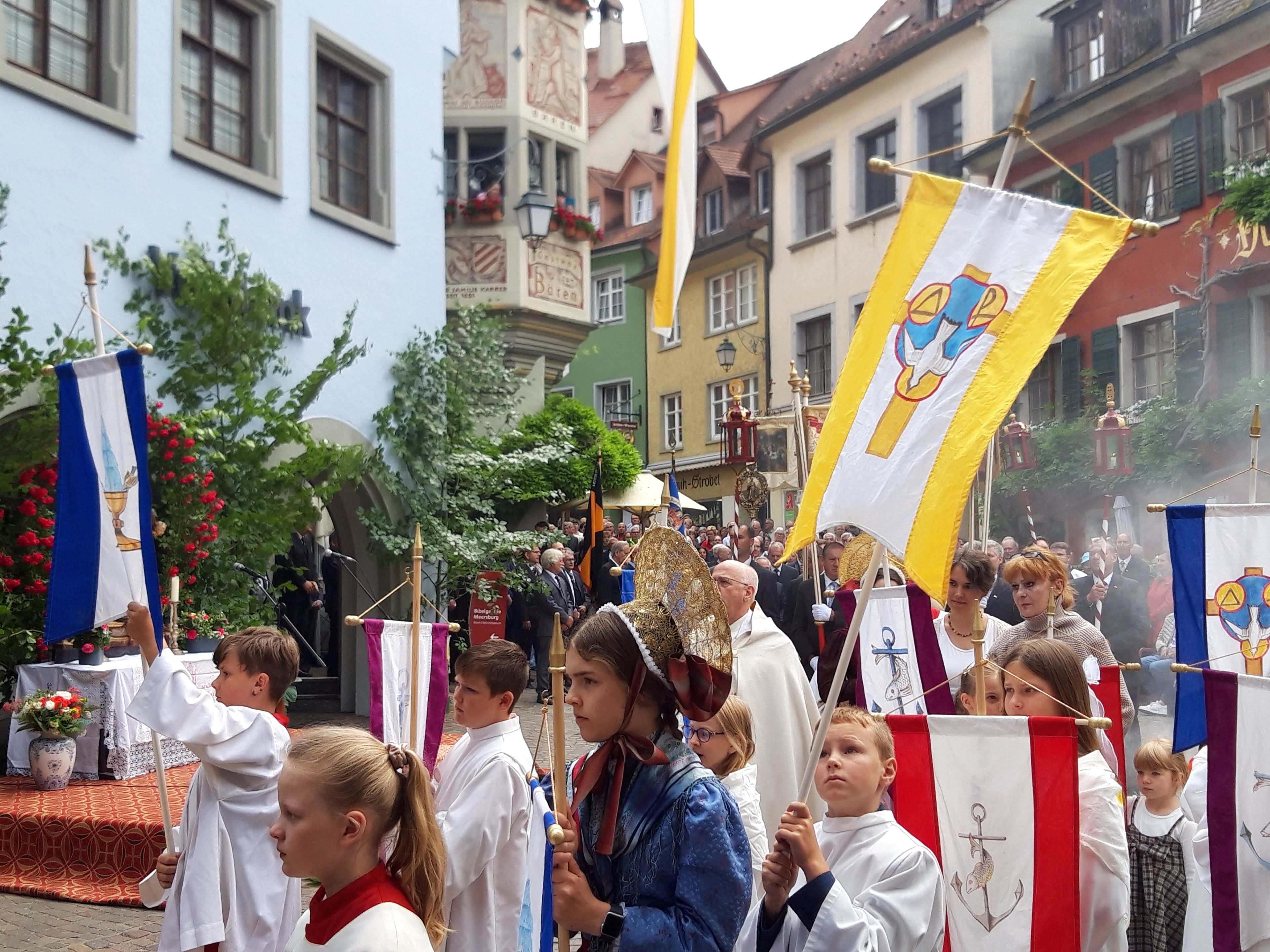 A group of people in traditional attire, holding flags and banners, in a street setting with buildings in the background.