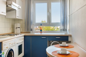 Una cocina con estufa, horno y lavadora, una ventana con cortinas, y una mesa con tazas y una cuchara.