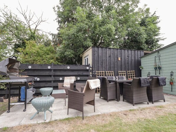 Een terras buiten met rieten meubels, een barbecue en een groene schuur op de achtergrond.