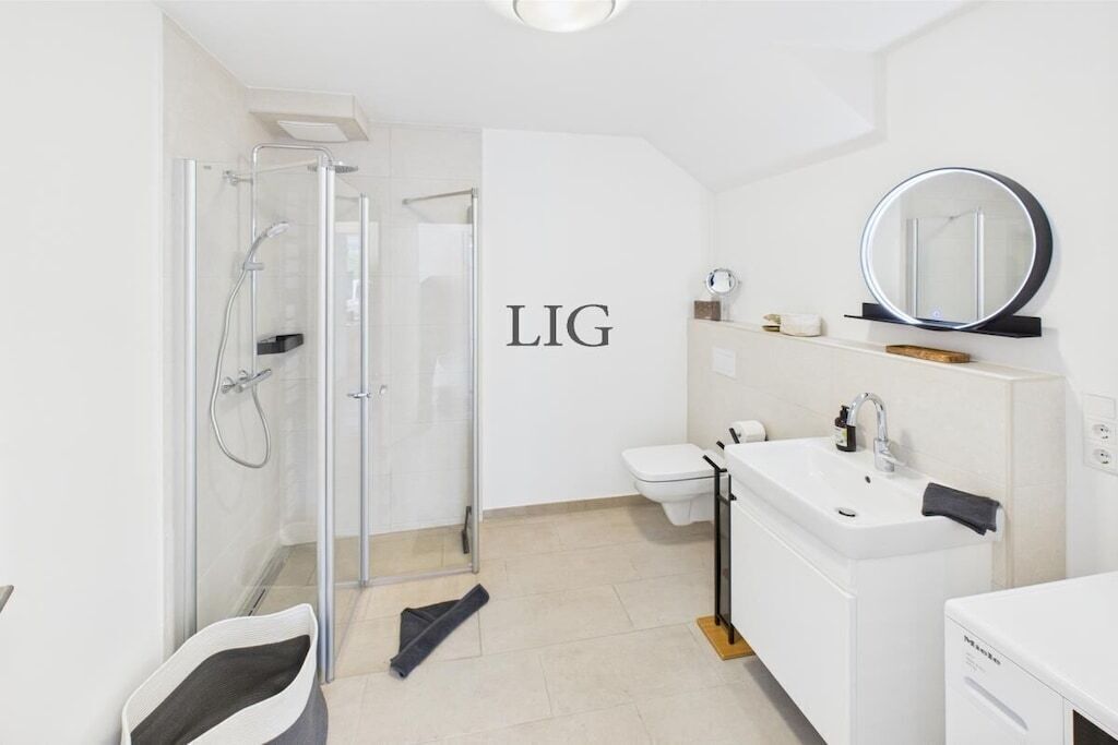 A modern bathroom with a glass shower enclosure, a toilet, a sink with a mirror, and a white wall with the text 'LIG'.