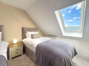 Ein Schlafzimmer mit schrägem Dach, einem Oberlicht, einem Bett mit grauer Decke, einem Nachttisch mit Lampe und einem Beistelltisch ebenfalls mit Lampe.