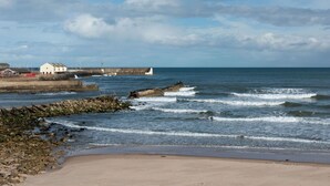 Uma paisagem costeira com praia de areia, linha de costa rochosa e um molhe que se estende para o mar.