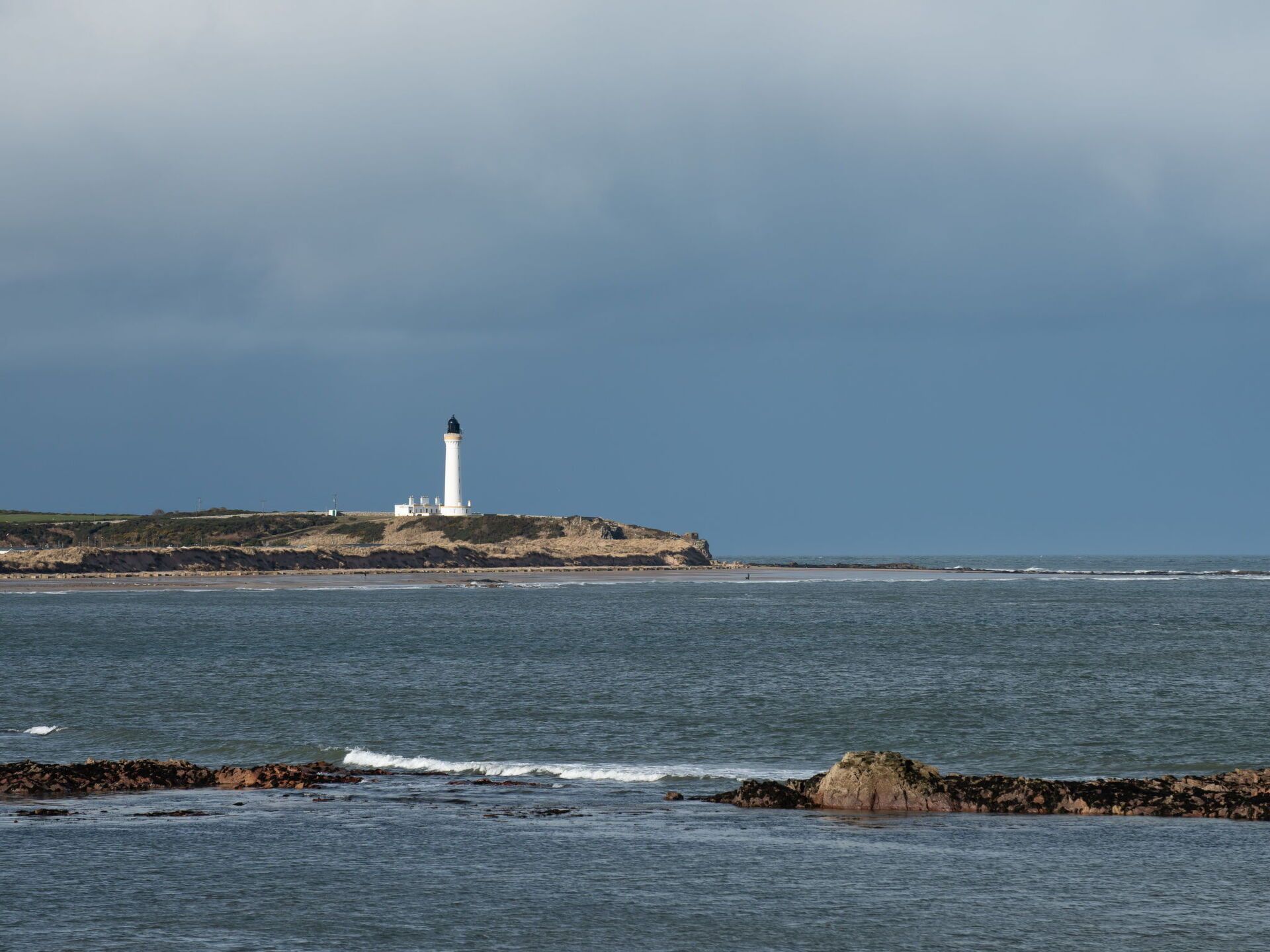 A lighthouse on a rocky coastline with calm sea and overcast sky.