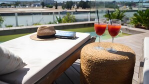 A table with a hat, a book, and two glasses of red drink, overlooking a body of water with a view of buildings and greenery. - Cozy Studio with All Amenities in One Space (El Puerto de Santa María)