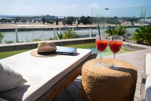 Une table sur laquelle se trouvent un chapeau, un livre et deux verres d’une boisson rouge. De la table, on a une vue sur une étendue d’eau, avec des bâtiments et de la verdure en arrière-plan.