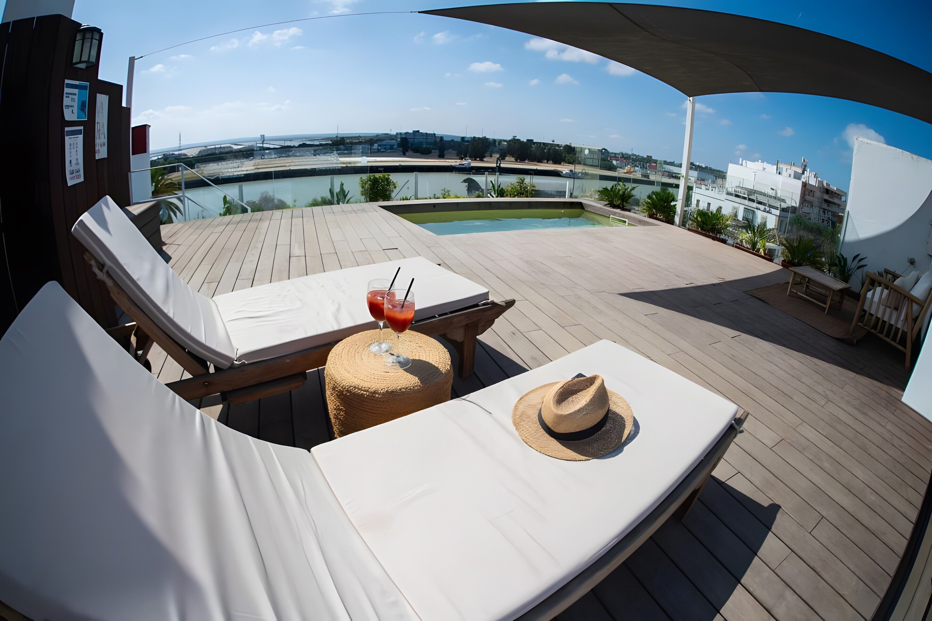 Une terrasse extérieure ensoleillée, dotée de chaises longues, d’une table avec des boissons, et offrant une vue sur une étendue d’eau et des bâtiments.