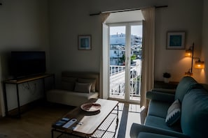 Een woonkamer met een bank, een salontafel met een schaal en een afstandsbediening, een televisie op een standaard en een balkon met een hekwerk.