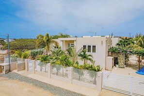 A modern two-story building with a flat roof, surrounded by a white fence and lush greenery. - Spacious Turibana villa with 4 king bedrooms, a private pool, tropical garden, palapa, BBQ, and separate apartment for added privacy. (Noord)