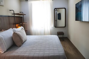 A neatly made bed with a quilted cover, a bedside table with a lamp, a mirror on the wall, and a window with curtains. - Bright, Well-Equipped Superior Apartment (El Puerto de Santa María)
