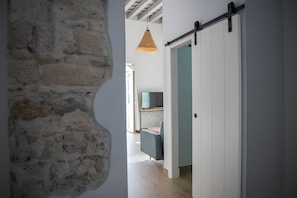 Een hal met een stenen muur, een schuifdeur van hout, een hanglamp en een blik op een kamer met een bureau en stoel.