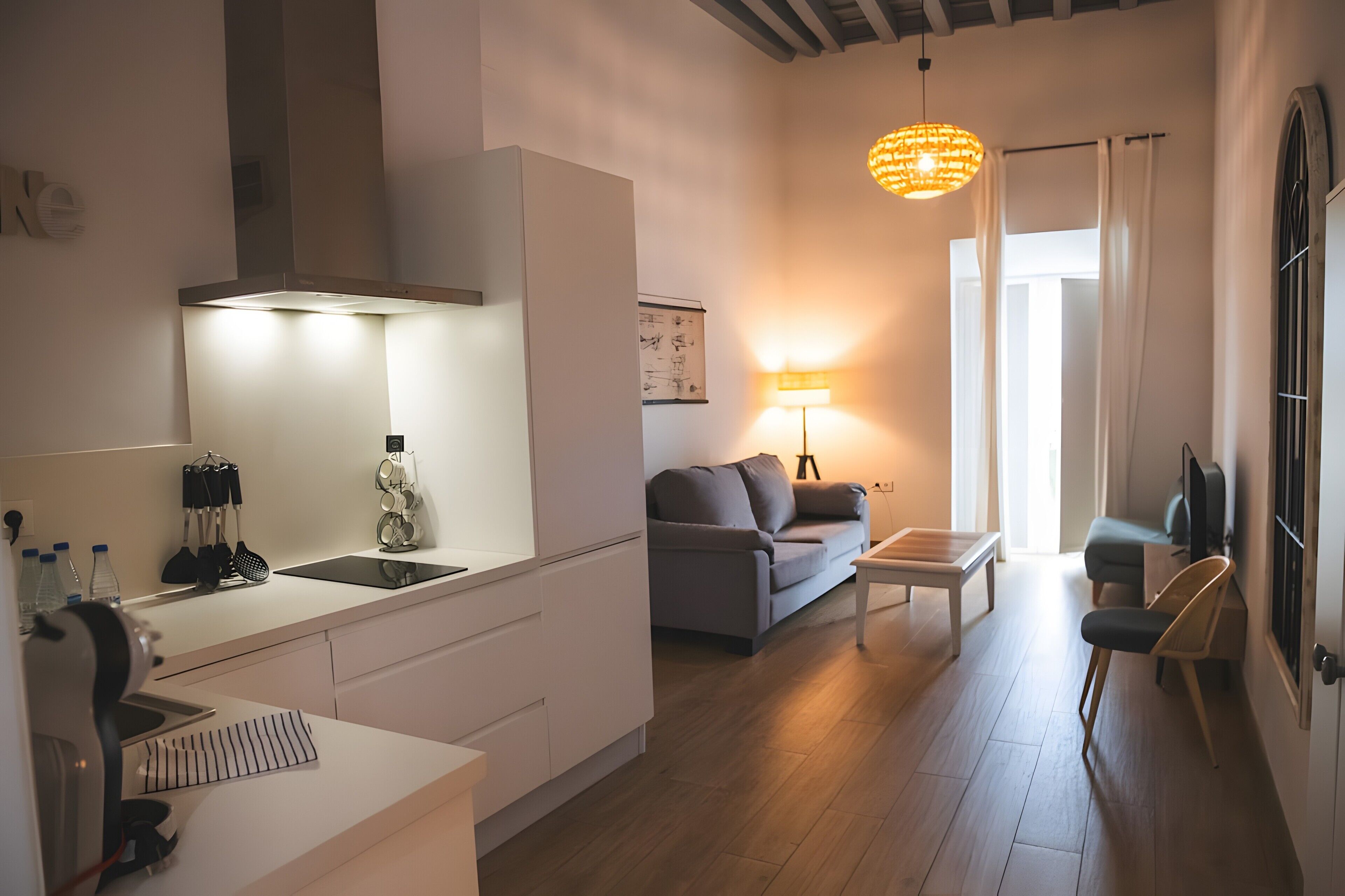 Eine moderne Küche mit Einbauherd, eine Essecke mit grauem Sofa und kleinem Tisch, sowie ein Wohnbereich mit grauem Sofa und Lampe.