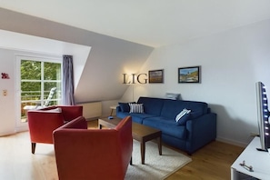 A modern living room with a blue sofa, red armchairs, a coffee table, and a large window with curtains. - Holiday apartment Island Love (Langeoog)