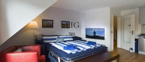 Une chambre avec un lit, une chaise rouge, une table en bois, un téléviseur et un coin cuisine.