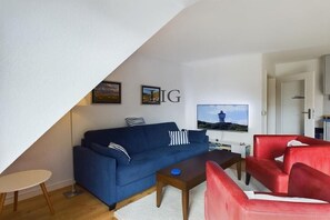 A living room with a blue sofa, a red armchair, a coffee table, and a TV on the wall. - Holiday apartment Island Love (Langeoog)
