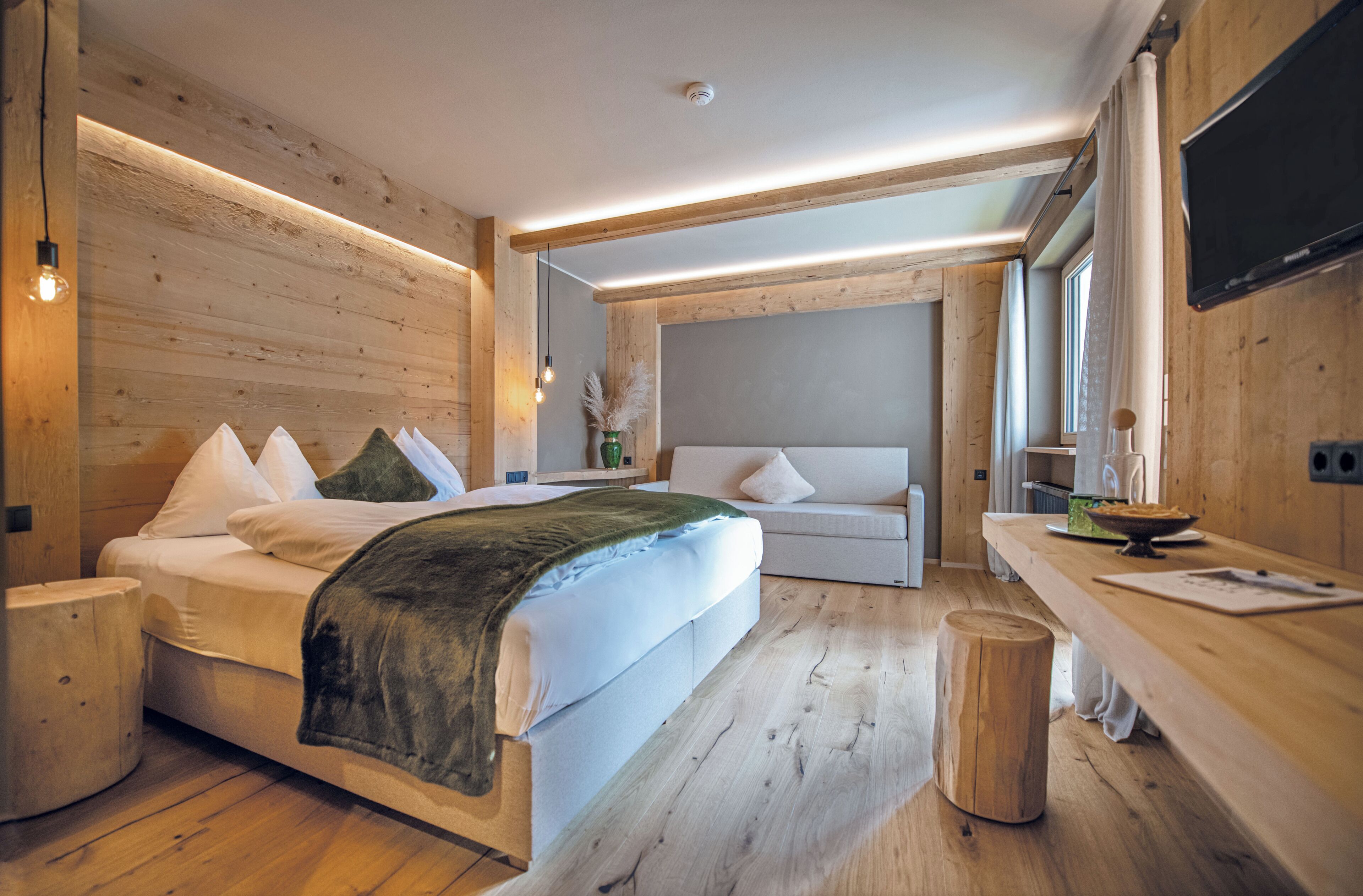 A modern bedroom with a large bed, a sofa, a wooden floor, and a flat-screen TV.