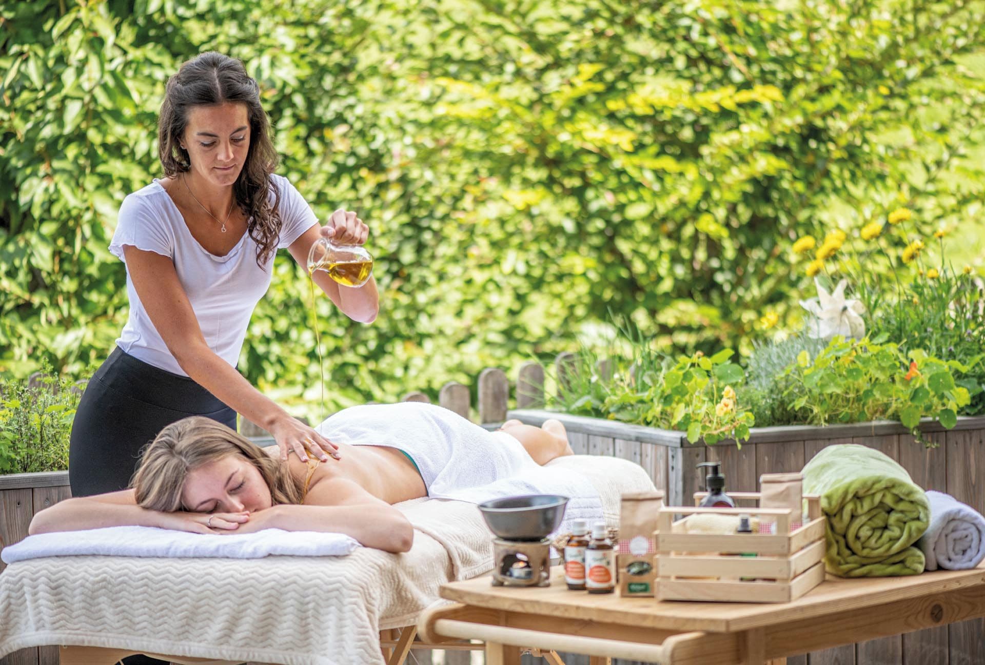 A person receiving a massage outdoors with a wooden table, a bowl, and bottles of oil.