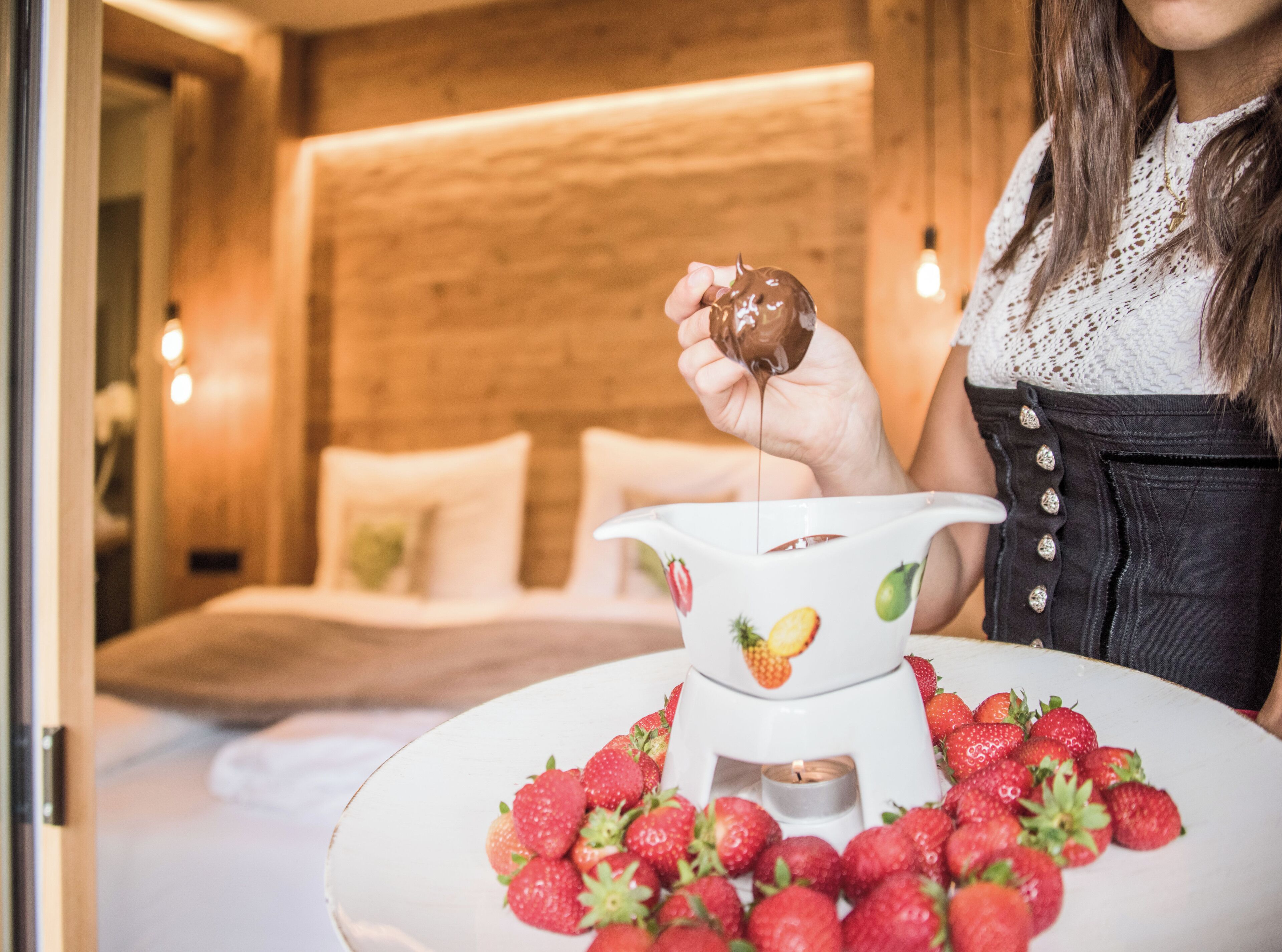 Una persona sostiene un dulce cubierto de chocolate sobre un plato de fresas, con una cama y una pared de madera al fondo.