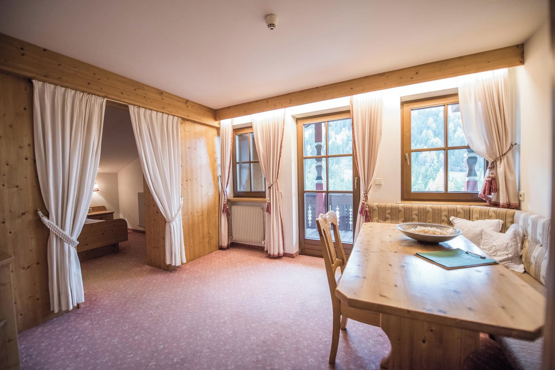 A room with wooden walls, a dining table with chairs, a window with curtains, and a radiator.