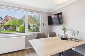 A modern room with a large window, a flat-screen TV mounted on the wall, and a dining table with chairs. - Erika holiday apartment (Langeoog)