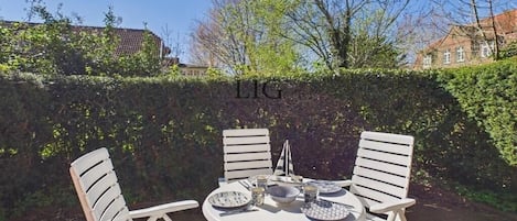Un espace de restauration en plein air avec une table ronde et des chaises blanches, entouré d’une haie.