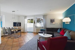 A modern living room with a sofa, a dining area with chairs, a TV, and a large window with curtains. - Holiday apartment Island Bliss (Langeoog)