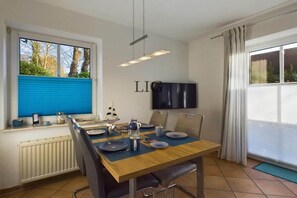 A dining area with a table set for two, a television mounted on the wall, and a window with blue blinds. - Holiday apartment Island Bliss (Langeoog)